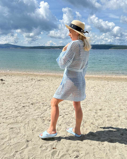 Model trägt ein blauen Langarm Cardigan, Einheitsgröße XS bis XL, Wickel mit Taillenbund, perfektes Strandkleid Knielänge, mit Lochmuster. Aus einer Mischung von 50 % Baumwolle und 50 % Acryl. Hintergrund ist ein Meer der kroatischen Küste der Adria. Nella Fashion Shop