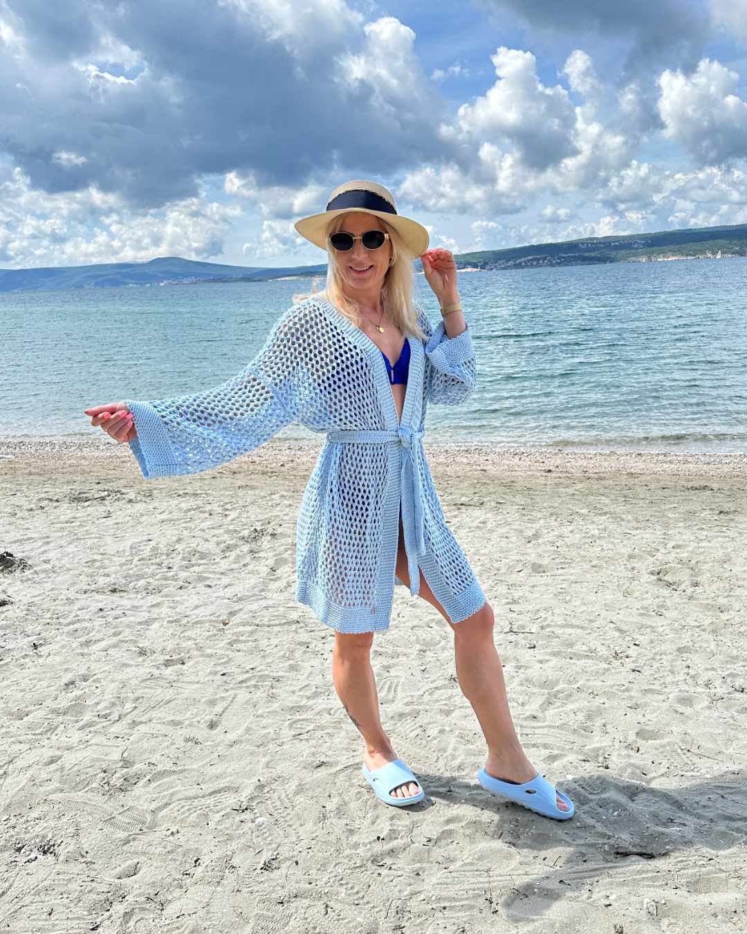 Model hat ein blauen Langarm Cardigan, Einheitsgröße XS bis XL, Wickel mit Taillenbund, perfektes Strandkleid Knielänge, mit Lochmuster. Aus einer Mischung von 50 % Baumwolle und 50 % Acryl. Hintergrund ist ein Meer der kroatischen Küste der Adria. Nella Fashion Shop