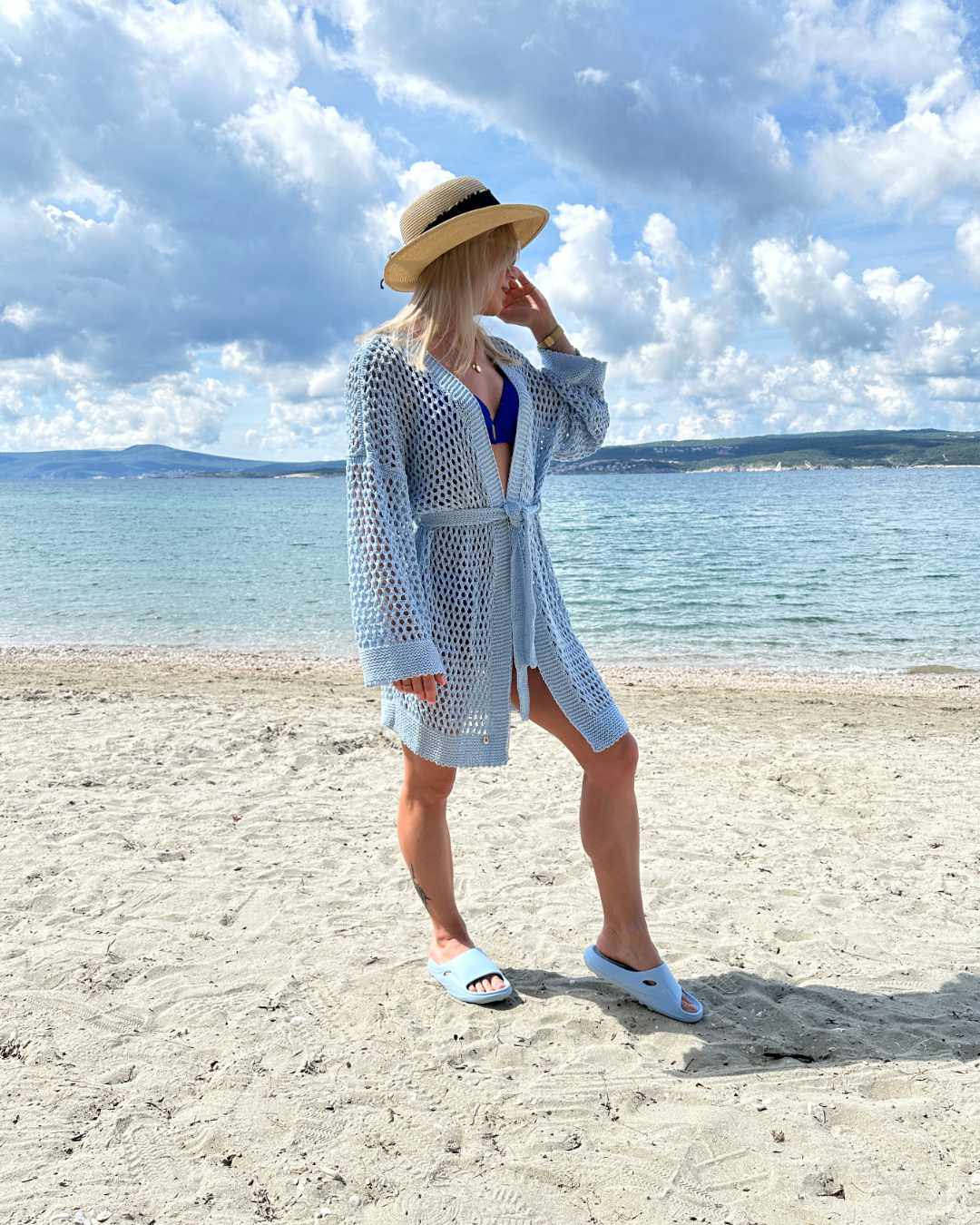 Model präsentiert ein blauen Langarm Cardigan, Einheitsgröße XS bis XL, Wickel mit Taillenbund, perfektes Strandkleid Knielänge, mit Lochmuster. Aus einer Mischung von 50 % Baumwolle und 50 % Acryl. Hintergrund ist ein Meer der kroatischen Küste der Adria. Nella Fashion Shop
