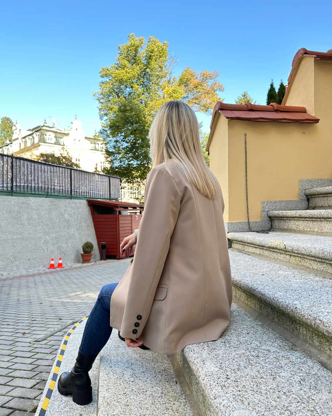 Model ist 160 cm groß und präsentiert von Hinten das Damen Doppelreihiger Oversized-Blazer lang mit Puffärmeln in Beige, mit Knöpfen verziert an den Manschetten. Blazer in der Größe 36 und 38 bei Nella Fashion Shop. Perfekter für Herbst oder Winter, mit Boots, Rollkragenpullover oder zu einem Kleid. Hintergrund sind Treppen zu Hotel.