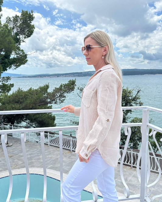 Model präsentiert den Langarm Damen Musselin Hemd aus 100 % Baumwolle in Beige. Leicht, locker und elegant Sommerhemd für Damen aus Musselin. In Oversized-Schnitt und Brusttasche. Erhältlich in XS/S und M/L, Schwarz und Beige bei Nella Fashion Shop. Hintergrund ist ein Meer der kroatischen Küste der Adria.