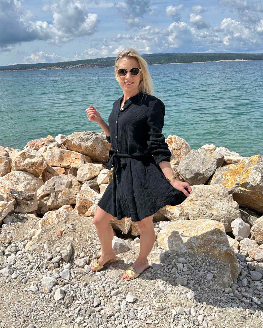 Model präsentiert das mini Damenmusselin-Kleid LUNA in Schwarz, Langarm Sommerkleid Musselin ist leicht locker elegant, Strandkleid LUNA ist vorne mit Knöpfen Verschluss, Hintergrund kroatisches Meer, Urlaub bei Nella & Fashion Shop