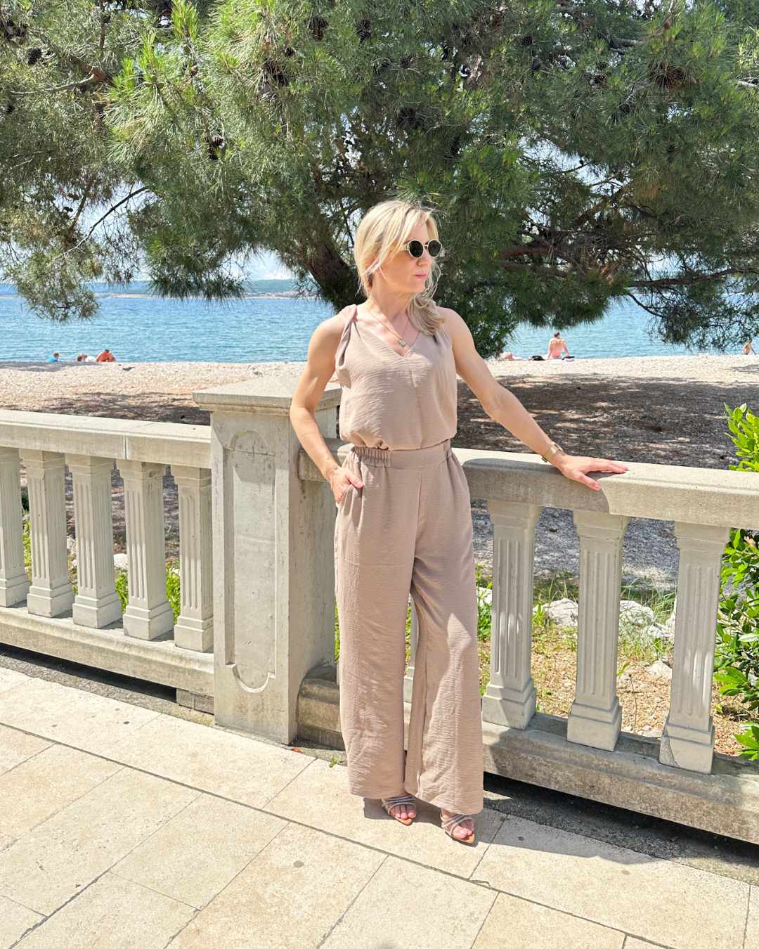Model präsentiert das Sommeroutfit Laura Damen Tank Top mit V-Ausschnitt plus Hosen mit weiten Beinen und hohem Bund zzgl. ein Gummi in der Taille in Beige Größe M/L. Alles auf eine kroatische Küste der Adria Hintergrund. Jetzt bei Nella Fashion Shop kaufen