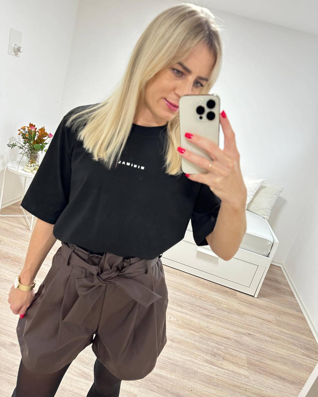 Model präsentiert die Damen Kunstleder Shorts mit hohem Bund in Schokoladenfarben Größe 34, mit einer Schleife/Gürte und Seitentaschen, kombiniert mit Schwarzen T-Shirt, perfektes Outfit für Frauen, auf Weißen Hintergrund, Nella Fashion Shop