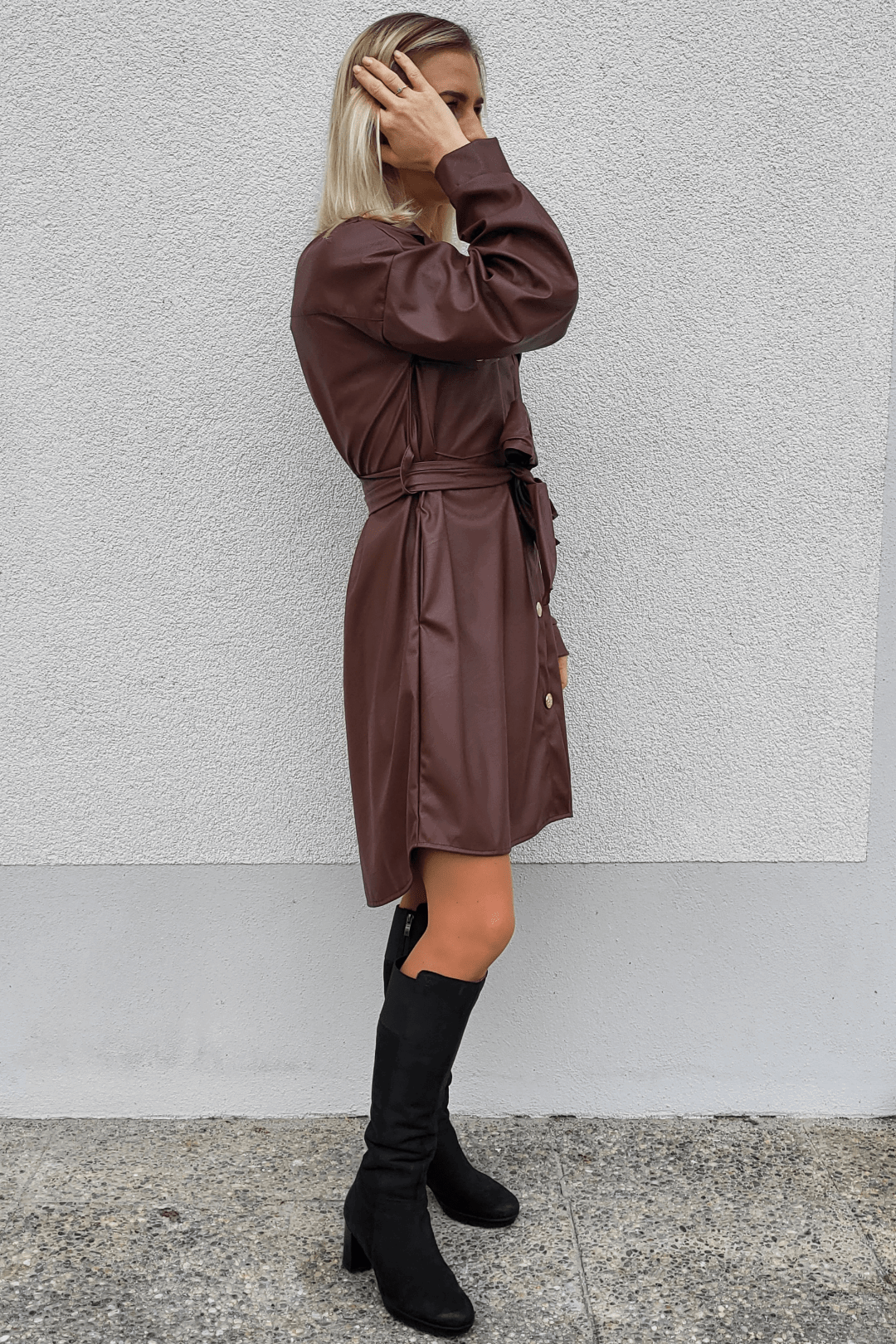 Model ist 160 cm groß und präsentiert an einer weißen Wand ein Herbst-Outfit mit Langarm Damen Kunstlederleid mit Knielänge in Schokoladen-Farbe dazu mit hohen Damenstiefeln Schwarz.