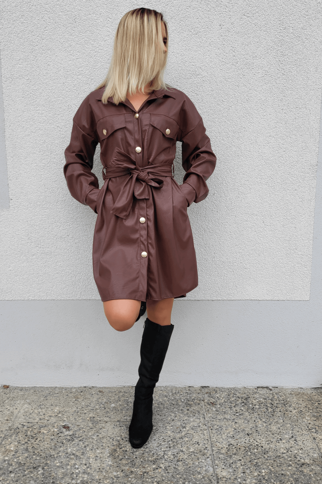 Model ist 160 cm groß und präsentiert an einer weißen Wand ein Herbst-Outfit mit Damen Kunstlederleid in Schokoladen-Farbe und V-Ausschnitt dazu, mit hohen Damenstiefeln Schwarz.