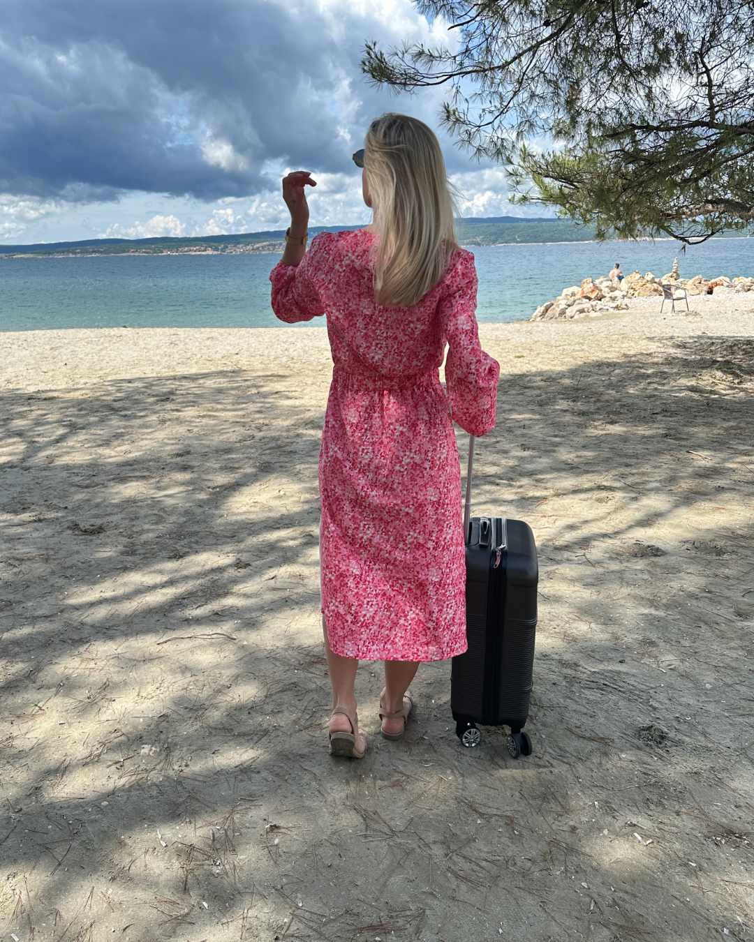 Model ist 160 cm groß und präsentiert von Hinten das S/M Knöchellanges elegantes Midi Langarm-Kleid in Rosa mit Weißen Punktenmuster, mit V-Ausschnitt, Seitenschlitz und elastischen Gummibündchen an den Armen sowie in der Teile, aus 60 % Viskose, 35 % Polyester, 5 % Elasthan, erhältlich in Größe S/M und L/XL, alles auf ein Meer Hintergrund mit ein Koffer für Urlaub, perfekt für Urlaub, Meer, Unterwegs, online kaufen bei Nella Fashion Shop