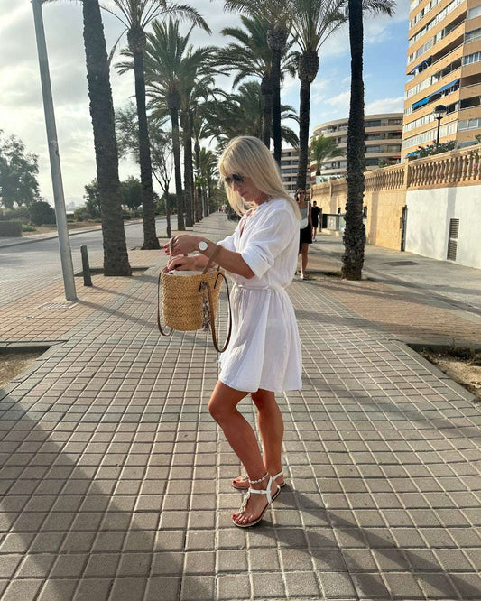 Model trägt das kurze mini Damen-Musselin-Kleid LUNA in weißer Farbe in der Mitte mit Schleife. Einen lässigen Schnitt. LUNA ist ein Sommerkleid aus Musselin 100 % Baumwolle. Vorne mit Verschluss auf Knöpfe. Leichtes, lockeres, elegantes Strandkleid oder Urlaubskleid, Hintergrund Palmen auf der Straße von Mallorca, Nella Fashion Shop