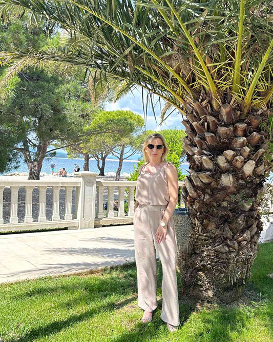 Model trägt das lockere zweiteilige Laura Sommeroutfit aus 97 % Polyester / 3 % Lycra: Tank Top mit tiefem Dekolleté plus Hosen mit weiten Beinen und hohem Bund in Beige, Größe M/L fällt zu groß aus. Alles unter eine Palme bei kroatische Küste der Adria Hintergrund. Nella Fashion Shop
