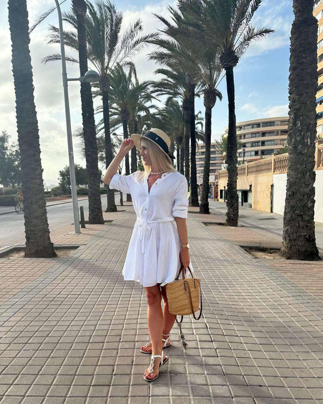 Die Dame trägt das weiße Musselin-Kleid in der Mitte mit Schleife. Einen lässigen Schnitt und V-Ausschnitt. LUNA ist ein Sommerkleid aus Musselin. Vorne mit Verschluss auf Knöpfe. Leichtes, lockeres, elegantes Strandkleid oder Urlaubskleid, Hintergrund Straße mit Palmen, Nella Fashion Shop
