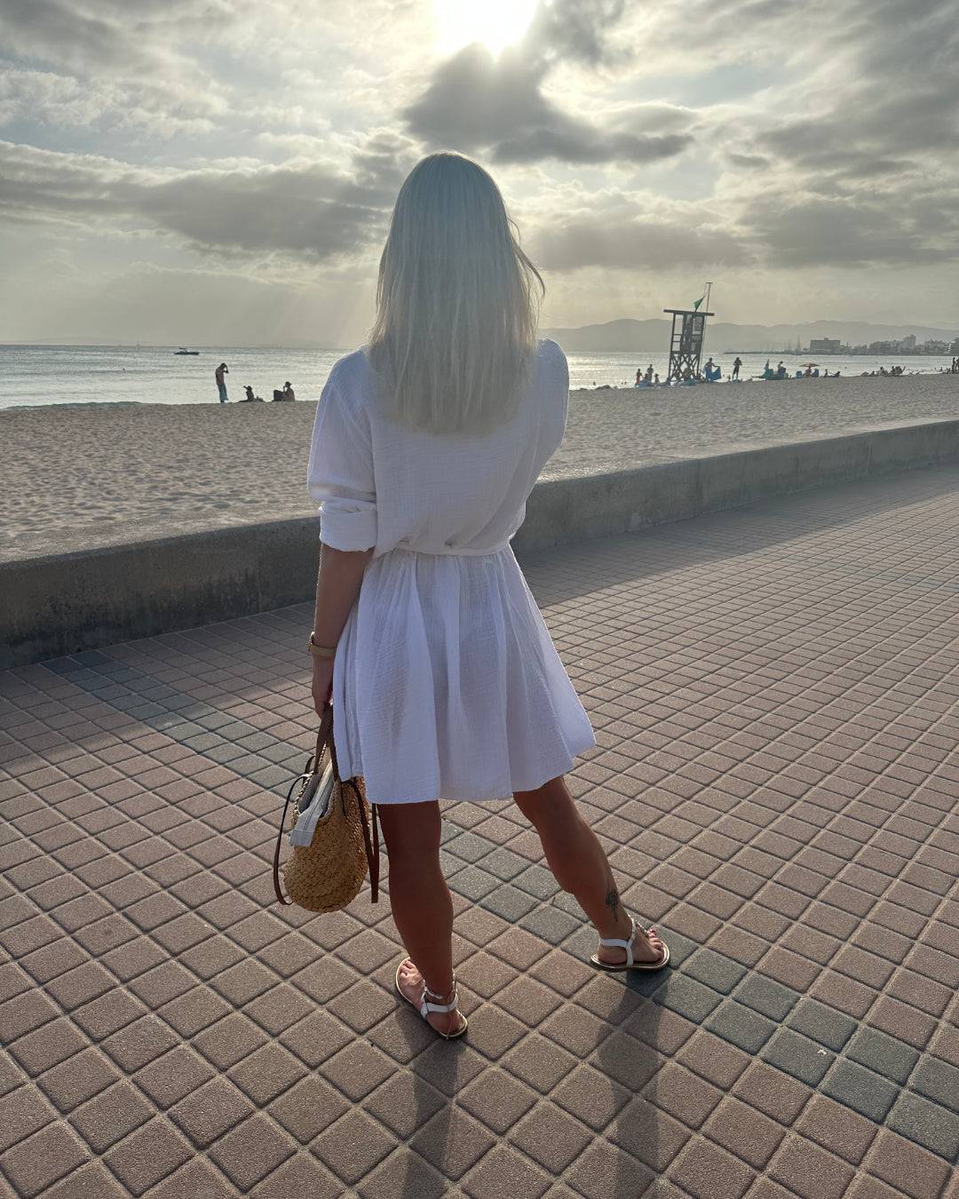 Dieses Model ist 160 cm und präsentiert von Hinten das Musselin Mini Kleid LUNA in weißer Farbe aus 100 % Baumwolle. Einen lässigen Schnitt. LUNA ist ein Sommerkleid aus Musselin. Vorne mit Verschluss auf Knöpfe. Leichtes, lockeres, elegantes Strandkleid oder Urlaubskleid, Hintergrund Meer von Mallorca, Nella & Fashion Shop
