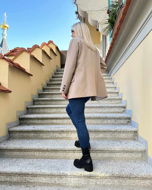 Model ist 160 cm groß und präsentiert von Hinten das Damen Doppelreihiger Oversized-Blazer lang mit Puffärmeln in Beige, mit Knöpfen verziert an den Manschetten. Blazer in der Größe 36 und  38 bei Nella Fashion Shop. Perfekter für Herbst oder Winter, mit Boots, Rollkragenpullover oder zu einem Kleid. Hintergrund sind Treppen zu Hotel. 