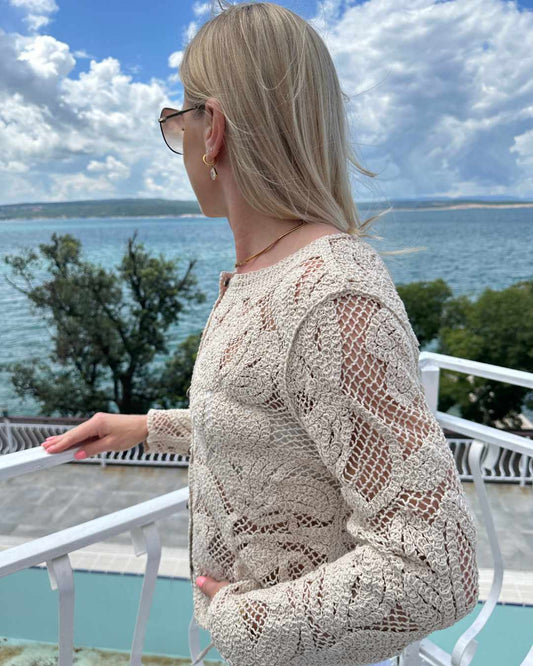 Model präsentiert von der Seite den Damen Boho Rundhals Strickpullover aus 100 % Baumwolle in Beige und Größe S/M. Strickart kleine Löcher und Muster Blumen. Hintergrund ist ein Meer der kroatischen Küste der Adria. Erhältlich bei Nella Fashion Shop in S/M oder M/L 