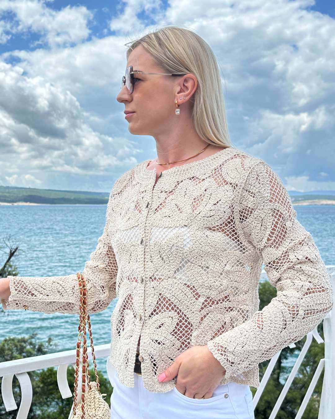 Model präsentiert von vorne den Damen Boho Rundhals Strickpullover aus 100 % Baumwolle in Beige und Größe S/M.  Holzknöpfe-Verschluss und die offene Strickart gibt Charmanten. Hintergrund ist ein Meer der kroatischen Küste der Adria. Erhältlich bei Nella Fashion Shop in S/M oder M/L 
