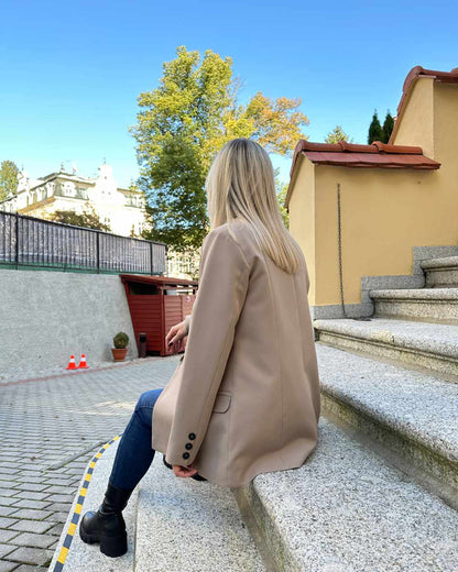 Model ist 160 cm groß und präsentiert von Hinten das Damen Doppelreihiger Oversized-Blazer lang mit Puffärmeln in Beige, mit Knöpfen verziert an den Manschetten. Blazer in der Größe 36 und  38 bei Nella Fashion Shop. Perfekter für Herbst oder Winter, mit Boots, Rollkragenpullover oder zu einem Kleid. Hintergrund sind Treppen zu Hotel. 