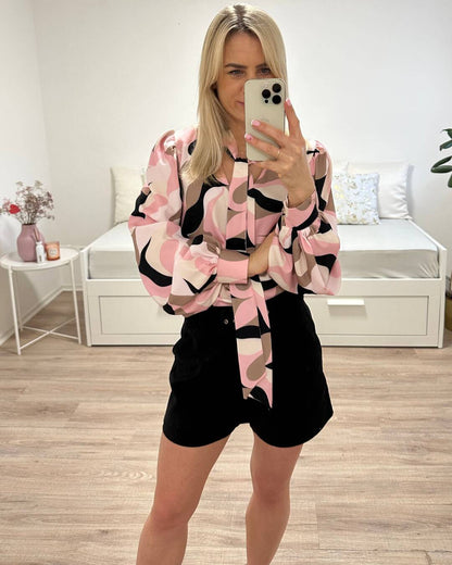 Model hat das Langarm-Hemd mit Schleife am Hals, somit kann eine Krawatte gemacht werden. Die Bluse ist mehrfarbig Abstract mit Rosa-Weiß-Schwarz und Braunen Farben. V-Ausschnitt und weiten Armen. Alles auf ein Weißenhintergrund, Nella Fashion Online Shop    