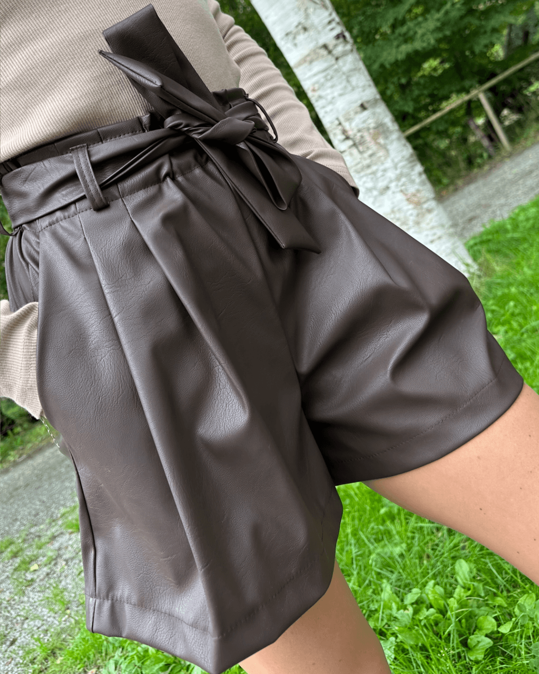 Das Model trägt die Damen Kunstleder Shorts Schokolade vorne gebunden in eine Schleife, Hände in Seitentaschen, Nella Fashion Shop 