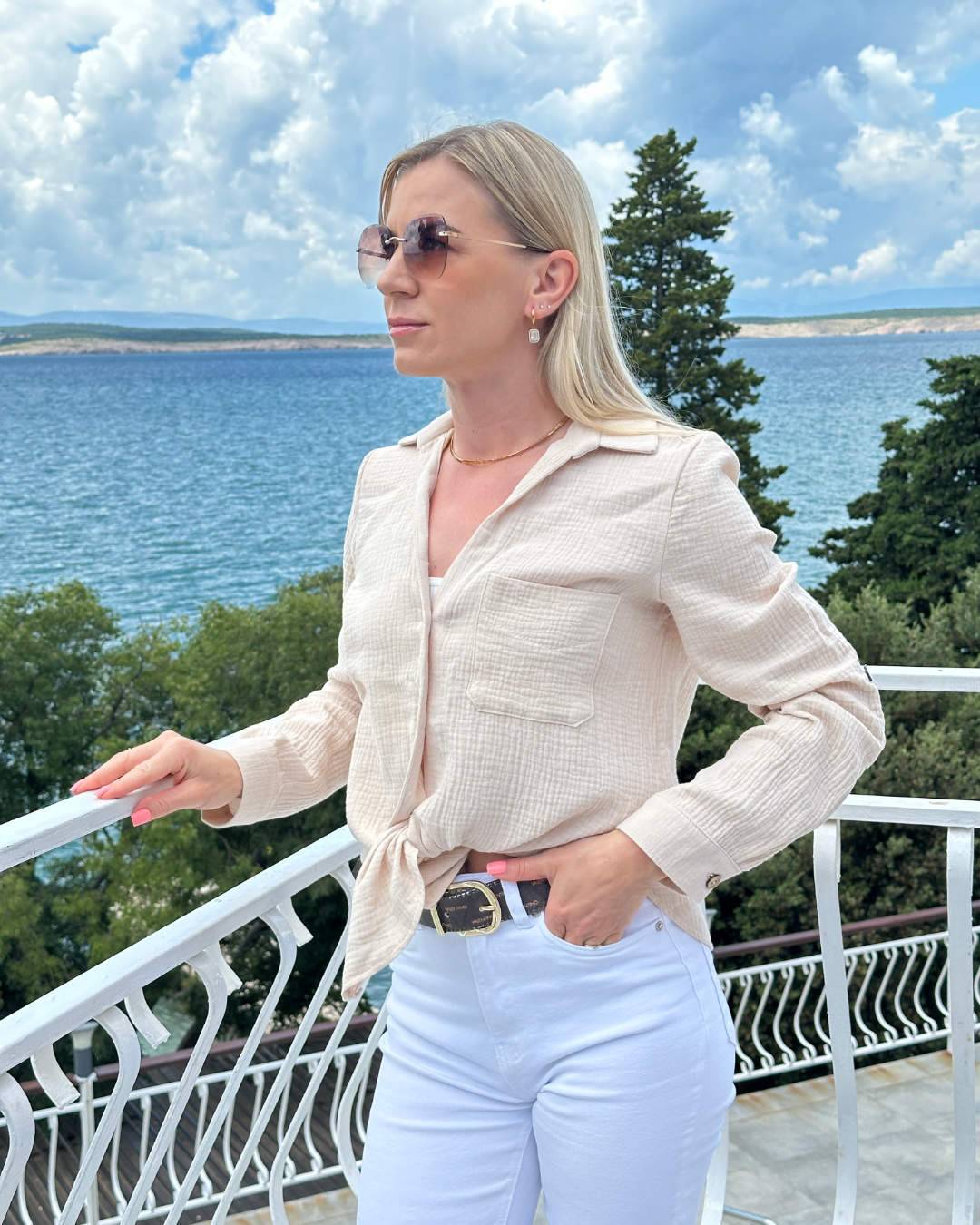 Model trägt Damen Musselin Hemd aus 100 % Baumwolle in Beige, leicht und luftig, sehr weiches Material.  Hemd mit Knöpfen und Brusttasche, Langarm mit Möglichkeit auf 3/4-Ärmeln und klassischem Umlegekragen in Oversized-Schnitt, dazu bequem und mit Stil. Frauenhemd mit V-Ausschnitt betont die Weiblichkeit. Hintergrund ist ein Meer der kroatischen Küste der Adria. Nella Fashion Shop