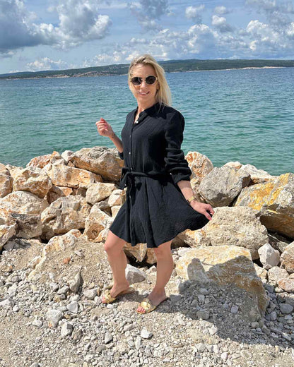 Model präsentiert das mini Damenmusselin-Kleid LUNA in Schwarz, Langarm Sommerkleid Musselin ist leicht locker elegant, Strandkleid LUNA ist vorne mit Knöpfen Verschluss, Hintergrund kroatisches Meer, Urlaub bei Nella & Fashion Shop