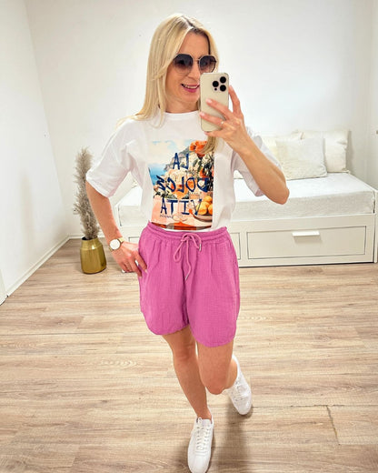 Model trägt das Weiße Rundhals T-Shirt mit Print (Aufdruck Beschreibung: Vordergrund: Schrift Al dolce vita, auf Tisch Orangen und Blumen, im Hintergrund Meer und ein Berg auf dem Hauser sind) aus 95 % Baumwolle / 5 % Elasta, Größe: Einheitsgröße XS bis L, aus ein Weißenhintergrund, Must-have für den Sommer, Accessoires: Handy und Brille, online Kaufen Nella Fashion Shop 
