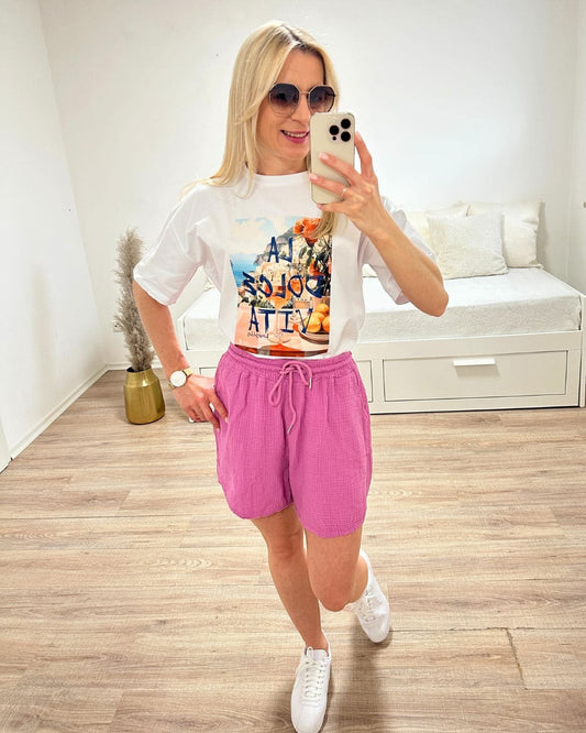 Model trägt das Weiße Rundhals T-Shirt mit Print (Aufdruck Beschreibung: Vordergrund: Schrift Al dolce vita, auf Tisch Orangen und Blumen, im Hintergrund Meer und ein Berg auf dem Hauser sind) aus 95 % Baumwolle / 5 % Elasta, Größe: Einheitsgröße XS bis L, aus ein Weißenhintergrund, Must-have für den Sommer, Accessoires: Handy und Brille, online Kaufen Nella Fashion Shop 
