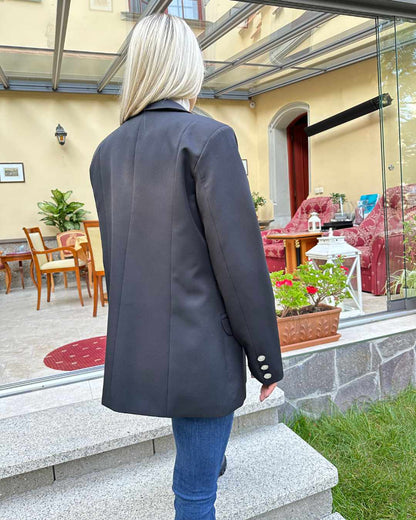 Model ist 160 cm groß und präsentiert von hinten ein Doppelreihiger Taillierter Damen-Blazer lang mit Puffärmeln regulärem Schnitt Schwarz in der Größe M. An den Manschetten mit 3 goldenen Knöpfen. Hintergrund Terrasse. Nella Fashion Shop 