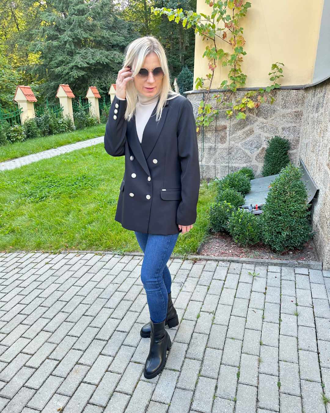 Model ist 160 cm groß und trägt ein Doppelreihiger taillierter schwarzer Damen-Blazer lang, mit Schulterposter, regulärem Schnitt und langen Ärmel in der Größe M. Aus Mischung 60 % Polyester / 35 % Viskose / 5 % Elastan. Der Blazer fällt zu groß aus. Perfekter für Herbst oder Winter, mit Boots, Rollkragenpullover oder zu einem Kleid. Hintergrund ist ein Hotel in Tschechien. Nella Fashion Shop
