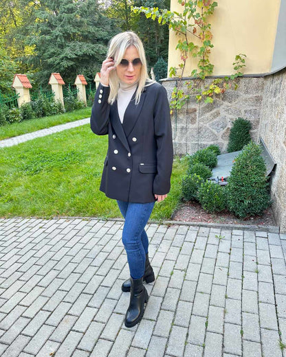 Model ist 160 cm groß und trägt ein Doppelreihiger taillierter schwarzer Damen-Blazer lang, mit Schulterposter, regulärem Schnitt und langen Ärmel in der Größe M. Aus Mischung 60 % Polyester / 35 % Viskose / 5 % Elastan. Der Blazer fällt zu groß aus. Perfekter für Herbst oder Winter, mit Boots, Rollkragenpullover oder zu einem Kleid. Hintergrund ist ein Hotel in Tschechien. Nella Fashion Shop