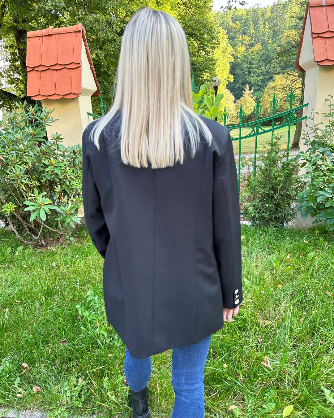 Model präsentiert ein Doppelreihiger Taillierter Damen-Blazer lang mit Puffärmeln regulärem Schnitt in der Größe M Schwarz von hinten. Fällt zu groß aus. Auf die Oberschenkel. Hintergrund Park. Nella Fashion Shop