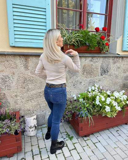 Model ist 160 cm groß und präsentiert das Damen Gerippter Rollkragenpullover Langarm in Hellbeige aus 95 % Baumwolle sehr weicher Stoff. Dazu blaue Jeans und Boots. Erhältlich bei Nella Fashion Shop in Größe M oder L.  Hintergrund Mauer mit Blumen und Fenster. 