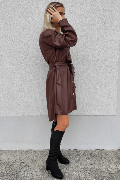 Model ist 160 cm groß und präsentiert an einer weißen Wand ein Herbst-Outfit mit Langarm Damen Kunstlederleid mit Knielänge in Schokoladen-Farbe dazu mit hohen Damenstiefeln Schwarz. 