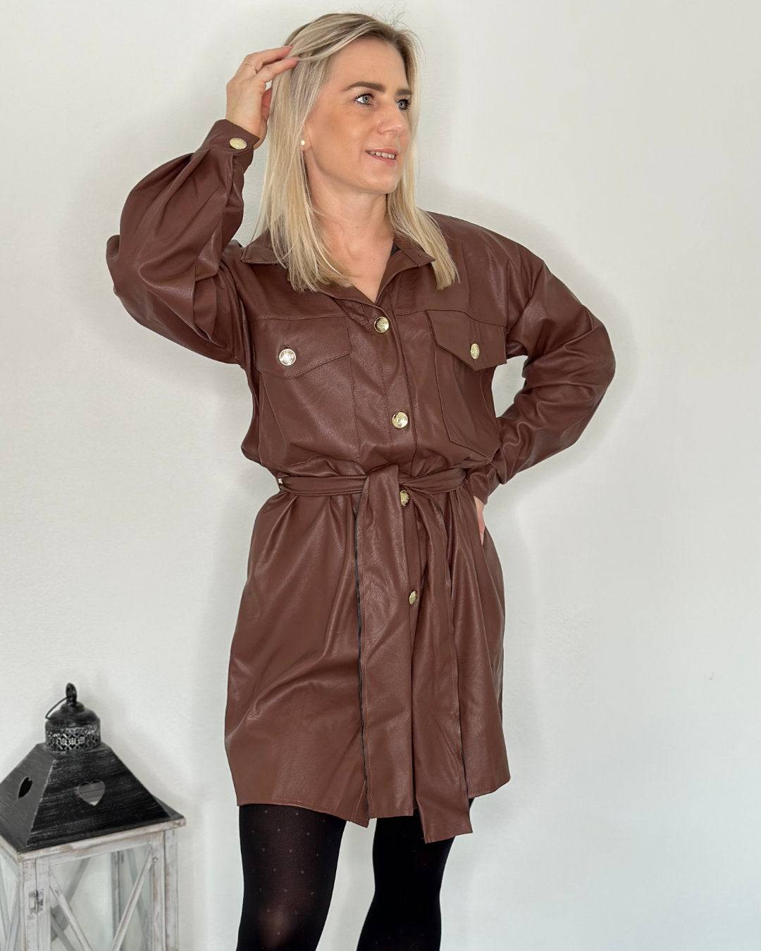 Model trägt auf ein Weißen-Hintergrund das Damen-Kunstlederkleid. Stil Eleganz mit Kragen, klassisch lange Ärmel und sportlich, mit zwei großen Seiten und Brusttaschen dazu goldenen Knöpfe Verschluss einen luxuriösen Akzent setzen. Gürtel gebunden in der Taille. Nella Fashion Shop 