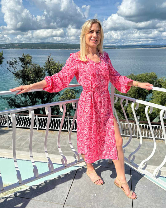 Model ist 160 cm groß und trägt das S/M Knöchellanges elegantes Midi Langarm-Kleid in Rosa mit Weißen Punktenmuster, mit V-Ausschnitt, Seitenschlitz und elastischen Gummibündchen an den Armen sowie in der Teile, aus 60 % Viskose, 35 % Polyester, 5 % Elasthan, erhältlich in Größe S/M und L/XL, alles auf ein Meer Hintergrund, perfekt für Urlaub, Meer, Unterwegs, online kaufen bei Nella Fashion Shop