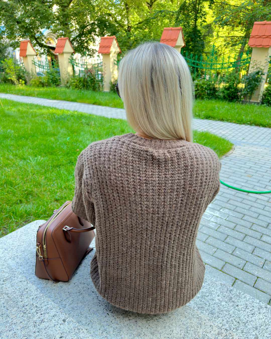 Model präsentiert im Park bei Sitzen von hinten den Oversized-Strickpullover Rundhals mit Zopfmuster Langarm Einheitsgröße XS/L in Schokoladenfarbe aus Materialien 15 % Mohair / 15 % Wolle / 10 % Polyester / 60 % Acryl. Erhältlich bei Nella Fashion Shop in Weiß, Dunkel beige, Schokoladenfarbe. 