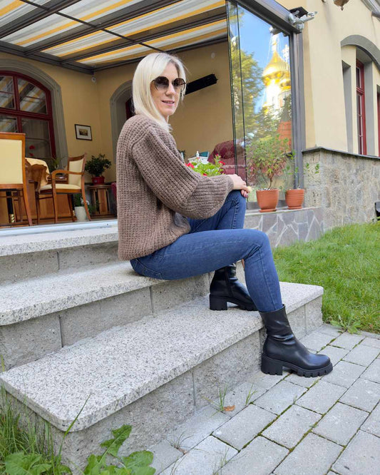 Model präsentiert Oversized-Strickpullover in Schokoladenfarbe mit einem wunderschönen Zopfmuster Langarm in der Einheitsgröße XS/L alles bei Sitzen auf ein Model präsentiert Oversized-Strickpullover in Weiß mit einem wunderschönen Zopfmuster Langarm in der Einheitsgröße XS/L alles bei Sitzen auf eine Terrasse. Nella Fashion Shop. 