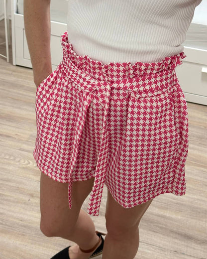 Model präsentiert auf weißen Hintergrund die Hoch taillierte Damen-Shorts mit Hahnentrittmuster in Einheitsgröße von XS bis L, mit elastischem Bund (Gummiband) in der Taille und einer Schleife zuzüglich Seitentaschen, Shorts mit Muster Rosa -Weiß. Nella Fashion Shop