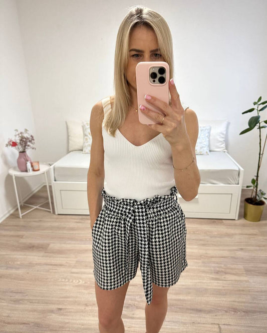 Model präsentiert auf weißen Hintergrund die Hoch taillierte Damen-Shorts mit Hahnentrittmuster in Einheitsgröße von XS bis L, mit elastischem Bund (Gummiband) in der Taille und einer Schleife zuzüglich Seitentaschen, Shorts mit Muster Schwarz -Weiß. Nella Fashion Shop