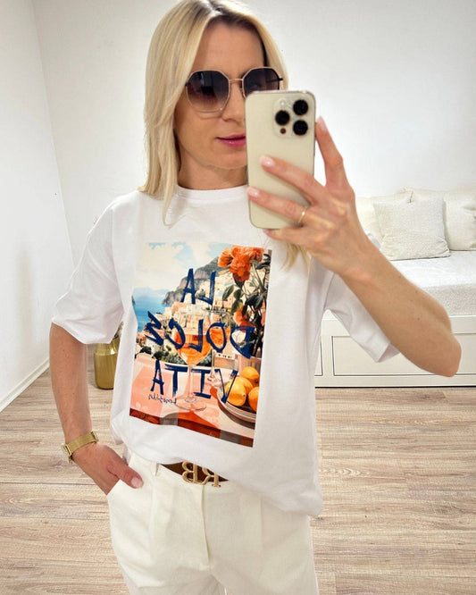 Model trägt das Weiße Rundhals T-Shirt mit Print (Aufdruck Beschreibung: Vordergrund: Schrift Al dolce vita, auf Tisch Orangen und Blumen, im Hintergrund Meer und ein Berg auf dem Hauser sind) aus 95 % Baumwolle / 5 % Elasta, Größe: Einheitsgröße XS bis L, aus ein Weißenhintergrund, Must-have für den Sommer, Accessoires: Handy und Brille, online Kaufen Nella Fashion Shop 
