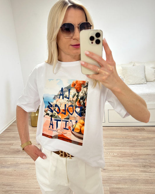 Model trägt das Weiße Rundhals T-Shirt mit Print (Aufdruck Beschreibung: Vordergrund: Schrift Al dolce vita, auf Tisch Orangen und Blumen, im Hintergrund Meer und ein Berg auf dem Hauser sind) aus 95 % Baumwolle / 5 % Elasta, Größe: Einheitsgröße XS bis L, aus ein Weißenhintergrund, Must-have für den Sommer, Accessoires: Handy und Brille, online Kaufen Nella Fashion Shop 

