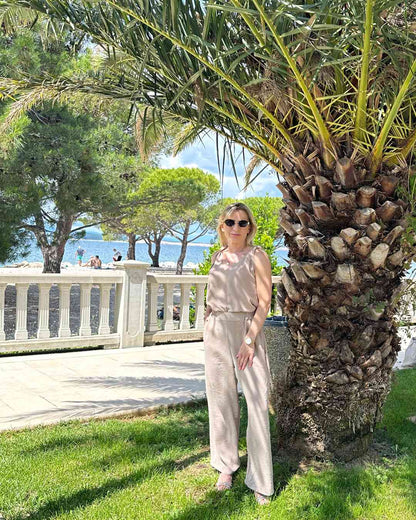 Model trägt das lockere zweiteilige Laura Sommeroutfit aus 97 % Polyester / 3 % Lycra: Tank Top mit tiefem Dekolleté plus Hosen mit weiten Beinen und hohem Bund in Beige, Größe M/L fällt zu groß aus.   Alles unter eine Palme bei kroatische Küste der Adria Hintergrund. Nella Fashion Shop 