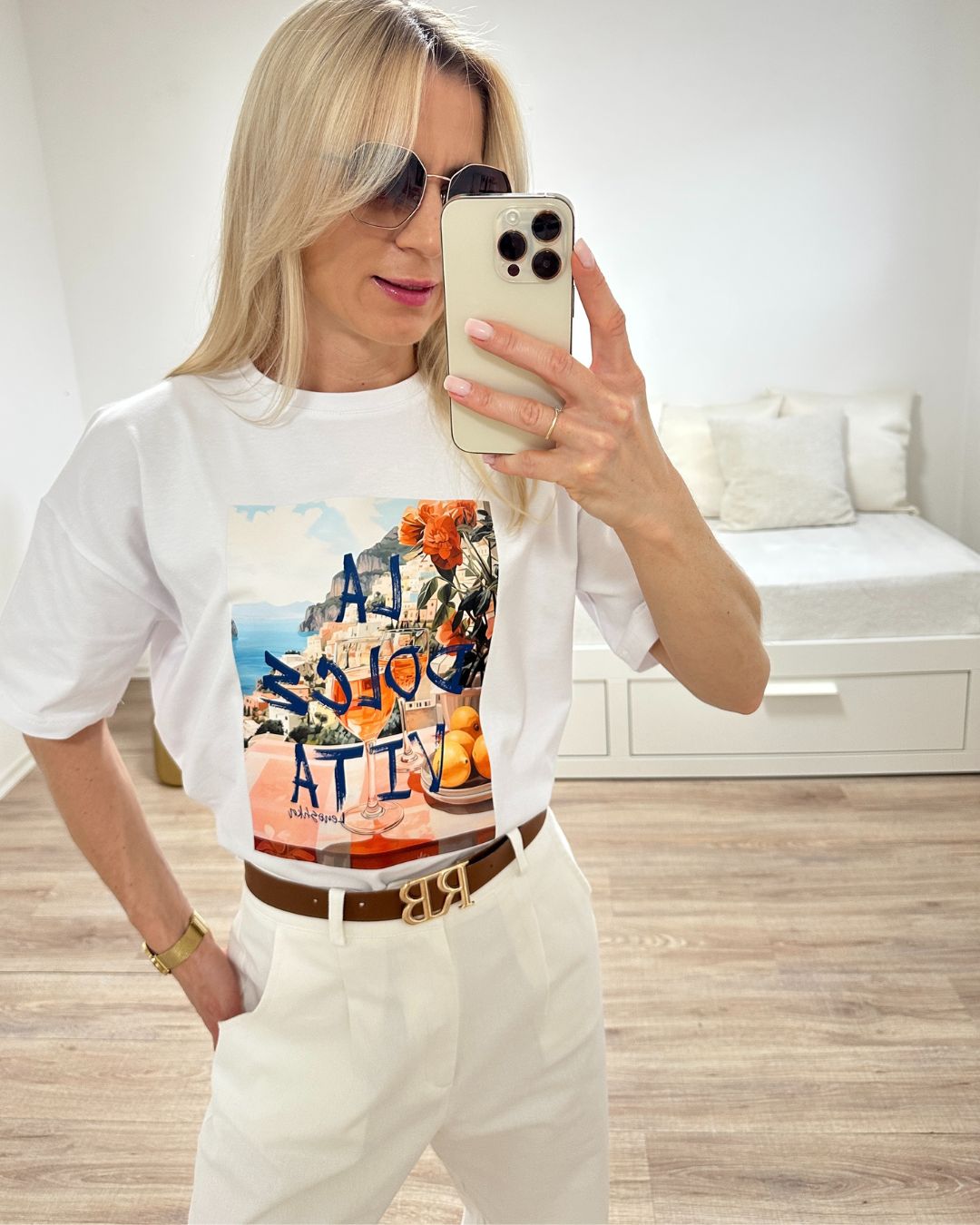 Model trägt das lockere Rundhals Oversized T-Shirt Damen in Weiß mit ein Print in der Mitte, dazu hat Sie Accessoires Brille, Handy, Armbanduhr und Gürtel, was ein perfekten-Look macht. Alles auf ein Weißenhintergrund, Nella Fashion Online Shop   
