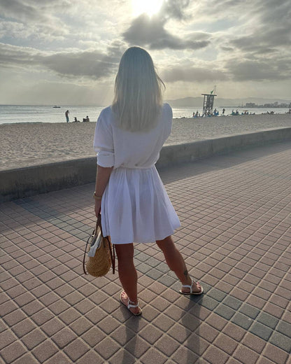 Dieses Model ist 160 cm und präsentiert von Hinten das Musselin Mini Kleid LUNA in weißer Farbe aus 100 % Baumwolle. Einen lässigen Schnitt. LUNA ist ein Sommerkleid aus Musselin. Vorne mit Verschluss auf Knöpfe.  Leichtes, lockeres, elegantes Strandkleid oder Urlaubskleid, Hintergrund Meer von Mallorca, Nella & Fashion Shop